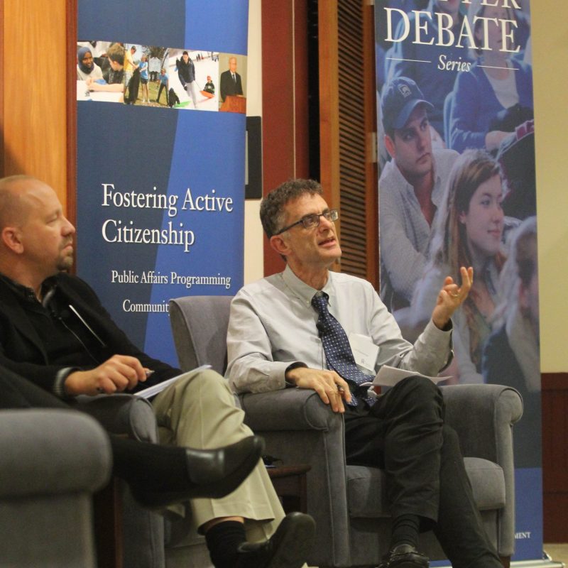 Two men seated on stage discussing civic engagement, with banners promoting active citizenship and a debate series in the background.