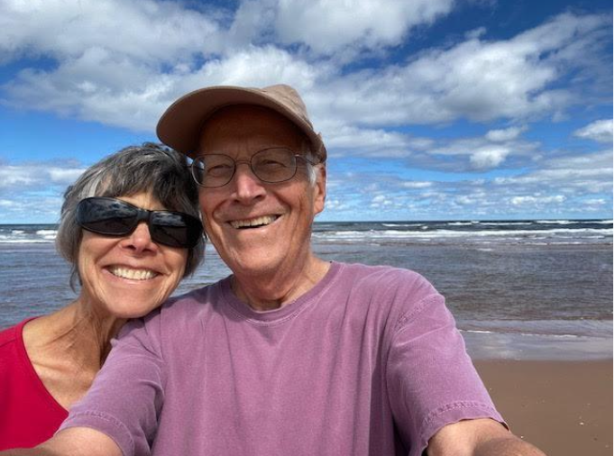A cheerful couple smiles for a selfie at the beach with a scenic ocean background and cloudy blue skies.