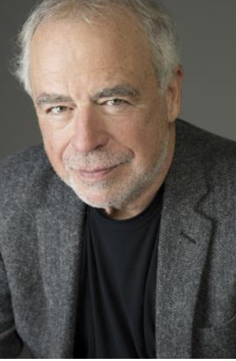 Older man with silver hair and beard, wearing a black shirt and gray blazer, smiling against a neutral background.