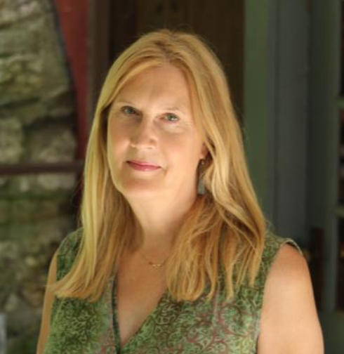 Woman with long blonde hair wearing a green dress, standing indoors with soft lighting and a natural background.