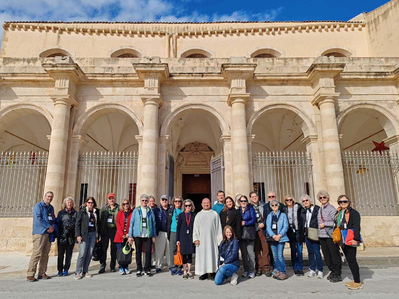 Group of tourists in front of historic building with arches, smiling in a sunny outdoor setting.