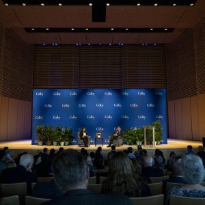 Discussion event in a modern auditorium with a blue backdrop featuring logos, audience seated, and speakers on stage.