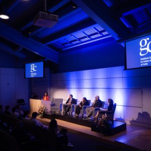 Panel discussion on stage at a center for public affairs, with a brightly lit backdrop and an engaged audience seated.