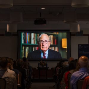 A speaker addresses an audience from a large screen in a conference room, with shelves of books visible behind him.