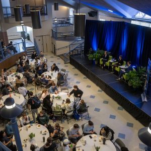A large gathering in a hall featuring tables with attendees and a stage with speakers illuminated by blue lights.