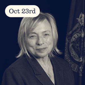 Portrait of a woman with short blonde hair in a suit, smiling against a dark background with a flag. Date displayed: Oct 23rd.