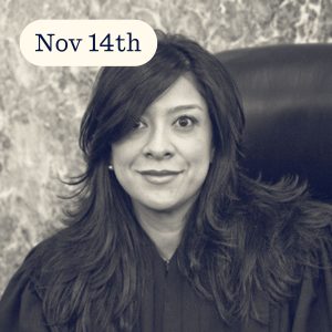 Portrait of a judge with long dark hair, wearing a black robe, seated in a courtroom with a date overlayed in the corner.