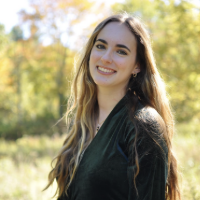 Young woman with long hair smiling in a sunlit outdoor setting with autumn foliage in the background.