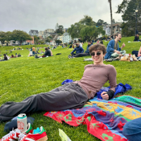 Person lounging on a colorful blanket in a park surrounded by other people enjoying a sunny day outdoors.