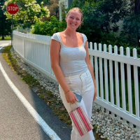 Smiling woman in a light top and white pants standing by a white picket fence near a stop sign on a sunny day.