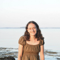 Young woman in a green dress stands by the water, smiling, with calm waves and a serene landscape in the background.