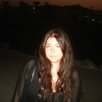 A woman with long dark hair poses outdoors at dusk, with buildings and palm trees in the background.