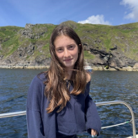 Smiling girl with long hair wearing a navy sweater stands on a boat with rocky shore and blue water in the background.