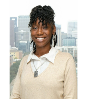 Smiling person with curly hair wearing a beige sweater in front of a city skyline.