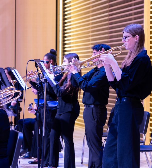 Musicians playing brass instruments on stage, wearing black attire, with a modern backdrop of wood paneling and lighting.