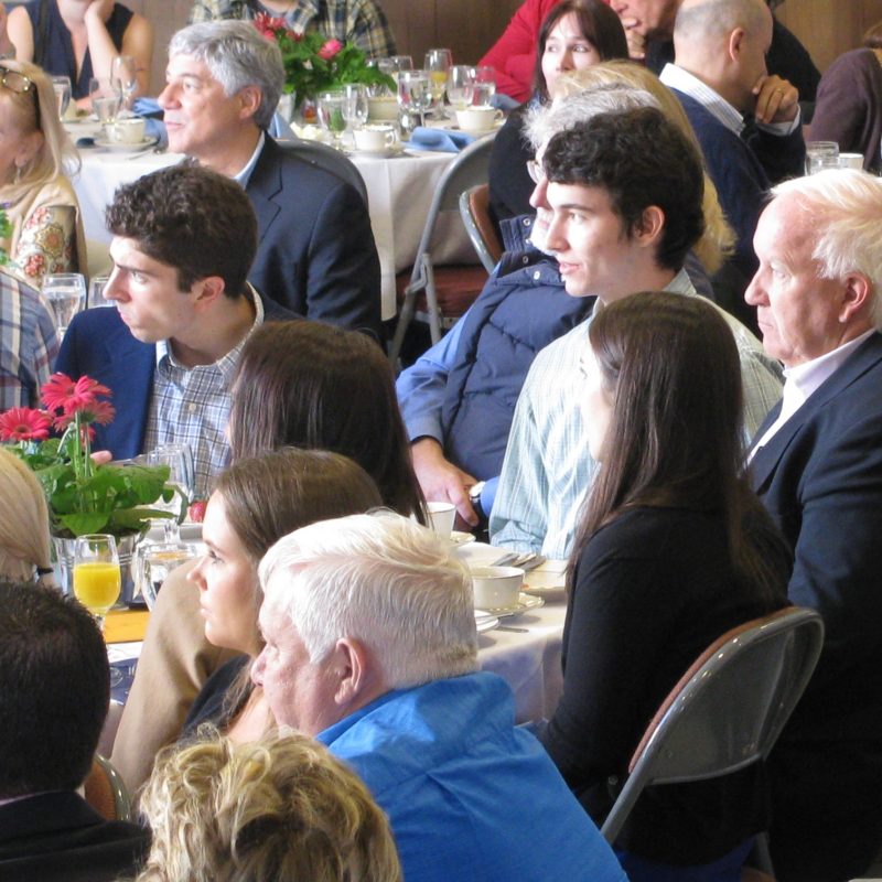 A group of people seated at a dining event, engaged and listening, with tables decorated with flowers and drinks.