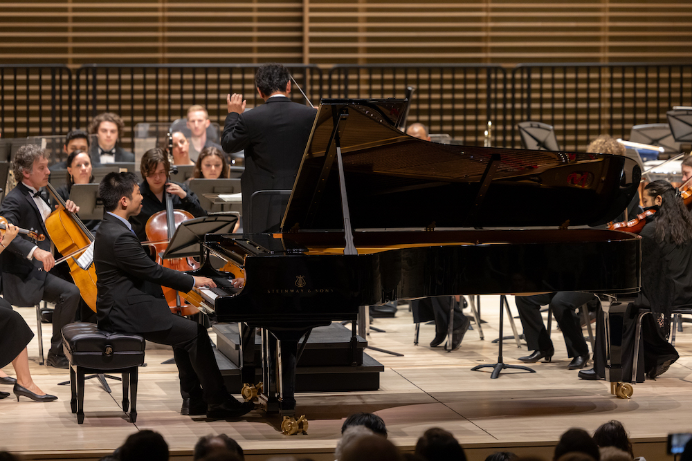 A solo pianist performs with an orchestra, featuring a grand piano and musicians in formal attire on stage.
