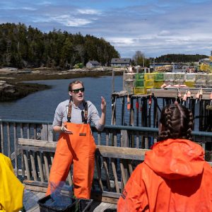 Person in orange overalls talks to two individuals in bright jackets near a dock with lobster traps and a scenic view.