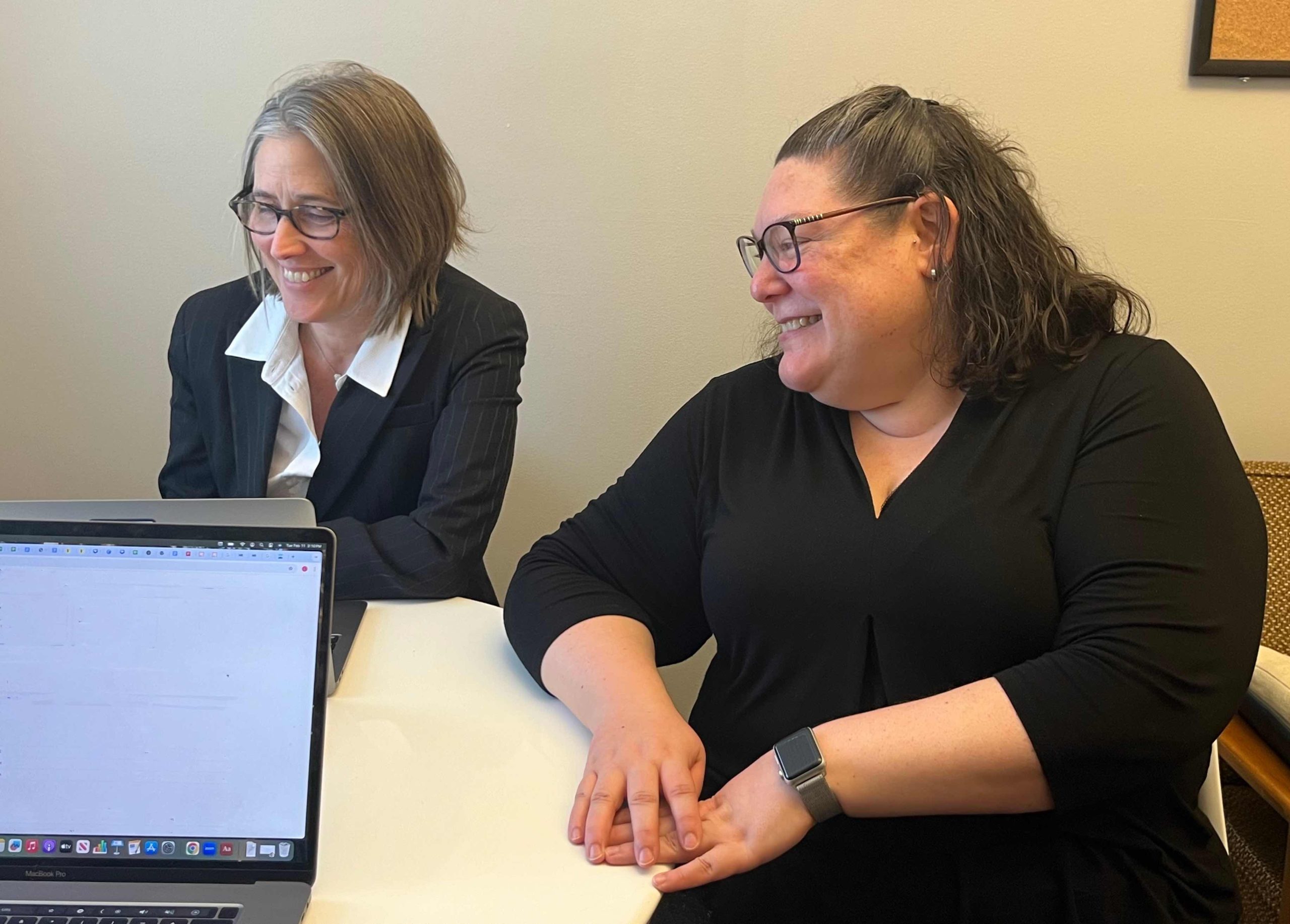 Two women in business attire sitting at a table, smiling and engaging with a laptop. Indoor setting with neutral colors.