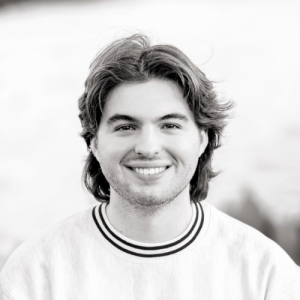 Smiling young man with medium-length hair and a light sweater against a blurred background of nature.