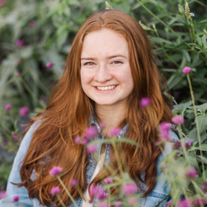 Smiling young woman with long red hair surrounded by green foliage and purple flowers. Bright and cheerful atmosphere.