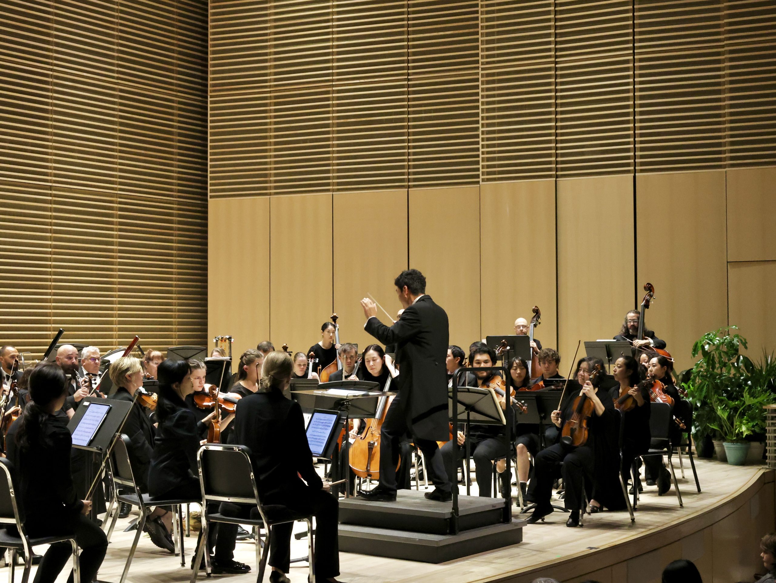 A conductor directs an orchestra performing on stage in a modern concert hall with musicians and instruments visible.