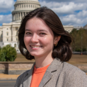 Young woman with shoulder-length dark hair smiling in front of a large building with a dome under a blue sky and clouds.