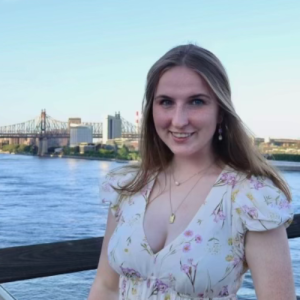 Young woman in a floral dress smiles by the water with a bridge and city skyline in the background under a clear blue sky.