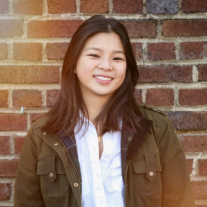 Smiling young woman in a green jacket stands in front of a brick wall with a warm, soft focus effect.