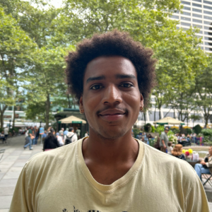 Young man with curly hair, wearing a light-colored shirt, stands in a park filled with people and green trees in the background.