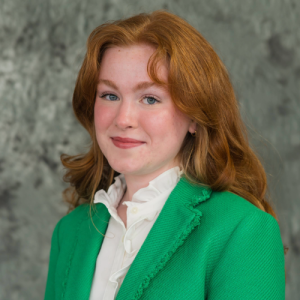 Young woman with red hair wearing a green blazer and white blouse, smiling against a textured gray background.