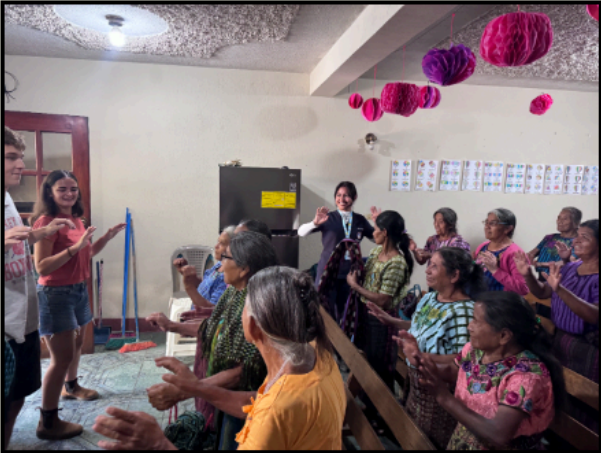 A group of elderly women joyfully participating in an activity, with a couple of young people engaging with them in a colorful room.