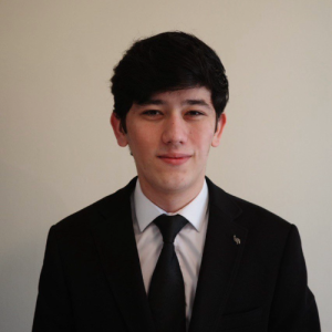 Young man in a black suit and tie, standing against a plain light background, with short dark hair and a neutral expression.