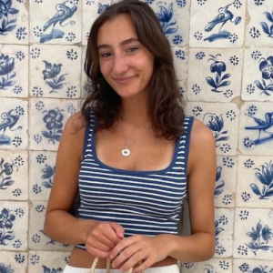 Young woman in a striped tank top smiles against a patterned blue and white tiled wall. She holds a small bag.