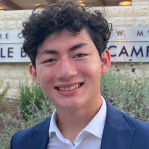 Young man with curly hair, wearing a suit jacket and white shirt, smiling in front of a building sign amidst greenery.