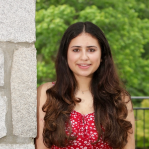 Young woman with long wavy hair smiles while wearing a red floral dress, standing near a stone wall and green foliage in the background.