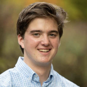 Young man with short brown hair smiling, wearing a light blue checked shirt against a blurred natural background.