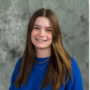 Portrait of a smiling young woman with long brown hair wearing a blue sweater and layered necklaces against a textured background.