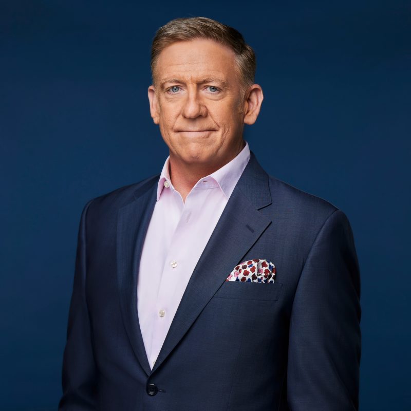 A man in a navy suit with a light pink shirt, standing confidently against a dark blue background. He has a patterned pocket square.