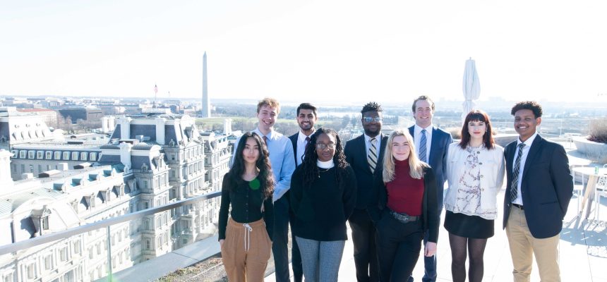 Group of diverse professionals posing on a rooftop with a city skyline and monument in the background on a sunny day.