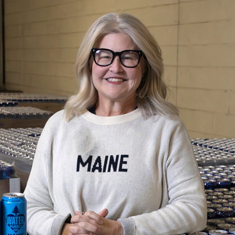 Woman with long blonde hair wearing glasses and a sweater that says MAINE, sitting in a warehouse with cans in the background.