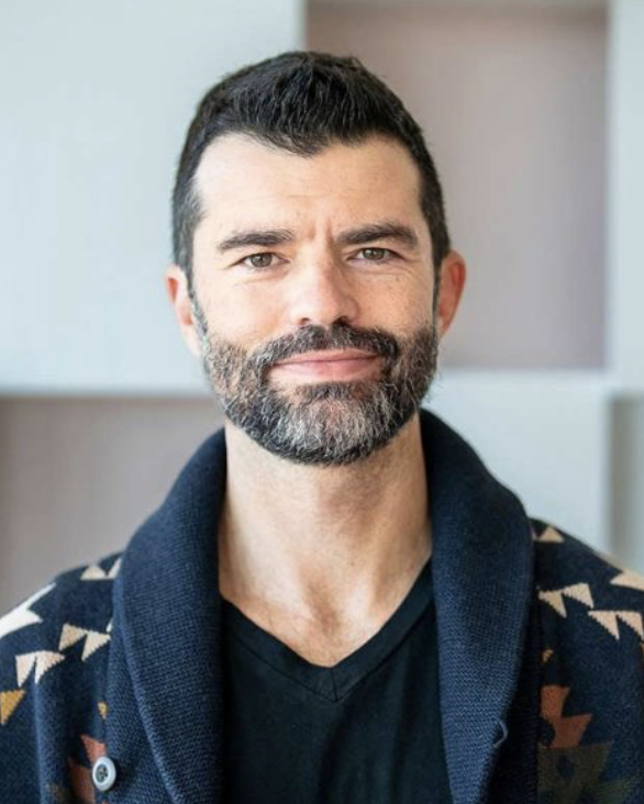 Smiling man with a beard wearing a dark sweater, posing against a light background with geometric shapes.
