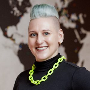 Person with short, light blue hair and a neon green chain necklace, smiling in front of a world map background.
