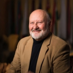 Smiling older man with a beard wearing a tan blazer, seated in a warmly lit environment with colorful backgrounds.