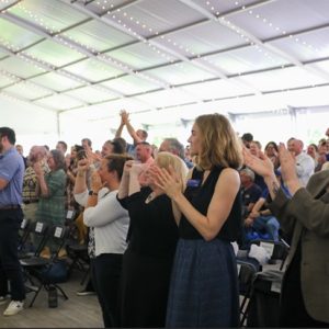 Crowd of people applauding under a large tent with string lights, showing excitement and celebration at an event.