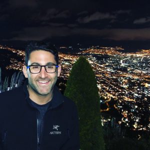 Man smiling with glasses against a night cityscape filled with lights and a green shrub in the foreground.