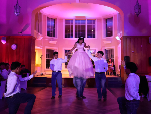 A young woman in a white gown is being lifted by friends on a dance floor, with colorful lights illuminating the background.