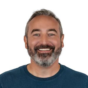 Smiling man with a beard wearing a blue shirt against a plain white background. Friendly and approachable expression.