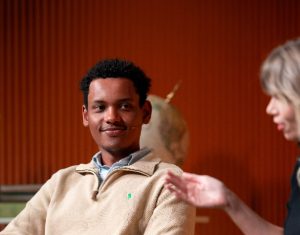 A young man smiles while listening to two women speaking in a warmly lit discussion setting. Green plant in background.