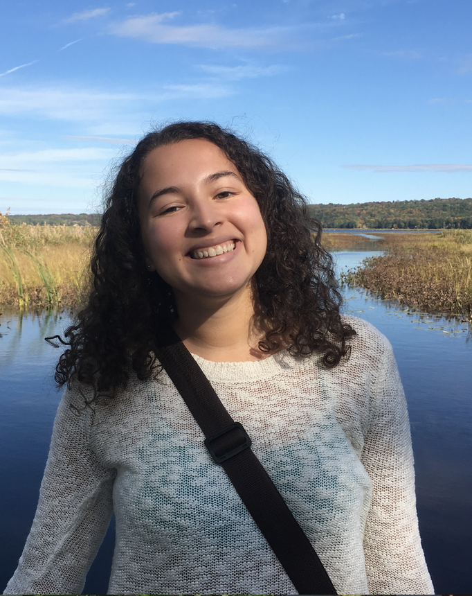 Smiling young woman with curly hair standing by a serene lake surrounded by greenery and blue sky.